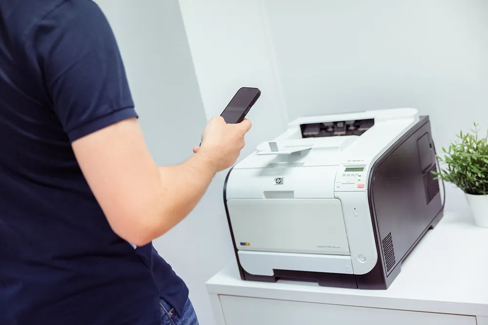 En som står med mobilen i hånden ved siden av en printer - Printix cloud print